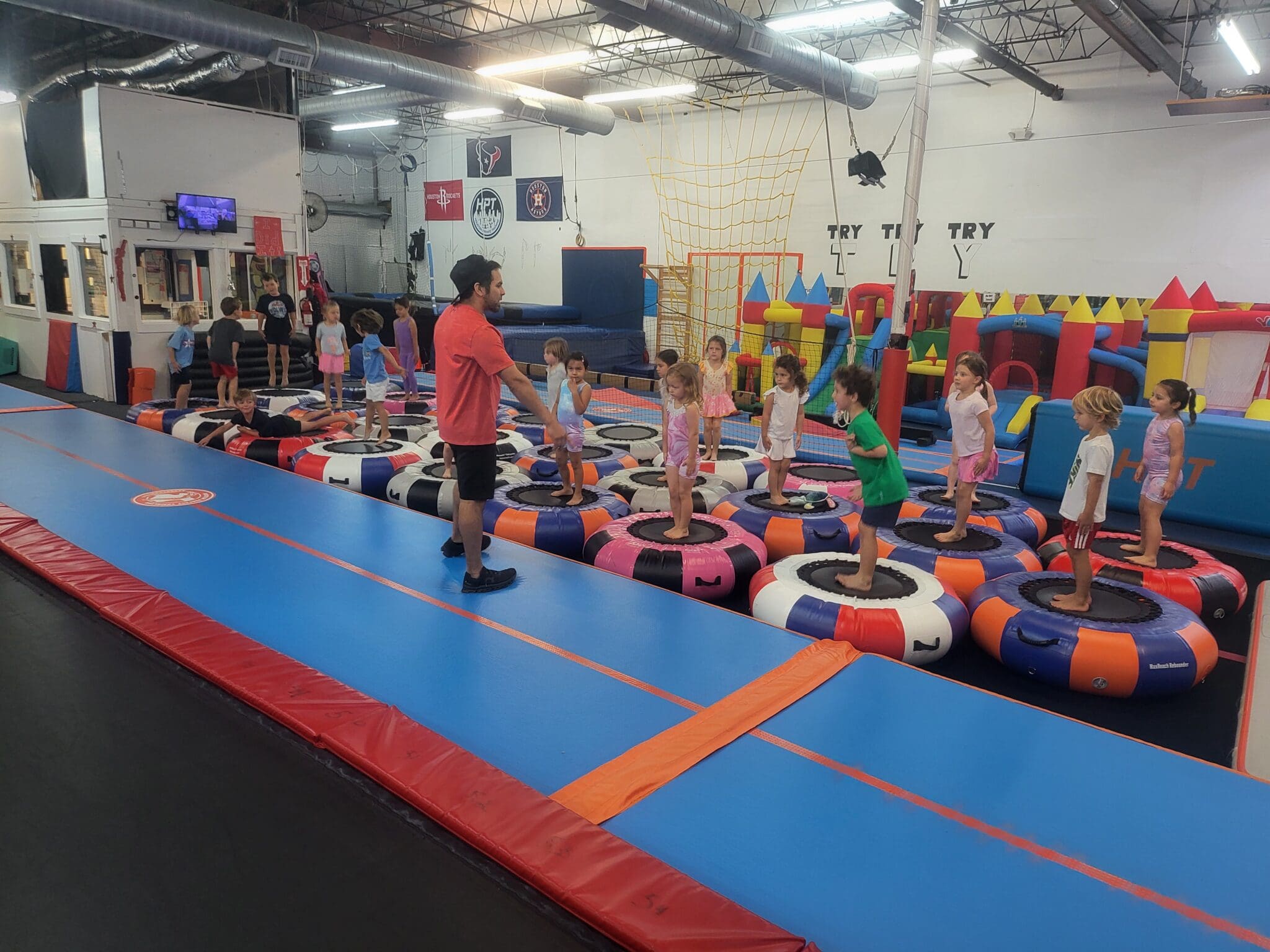 Children stand on trampolines in a gym, guided by an adult in a red shirt. Colorful equipment surrounds them in a bright space.