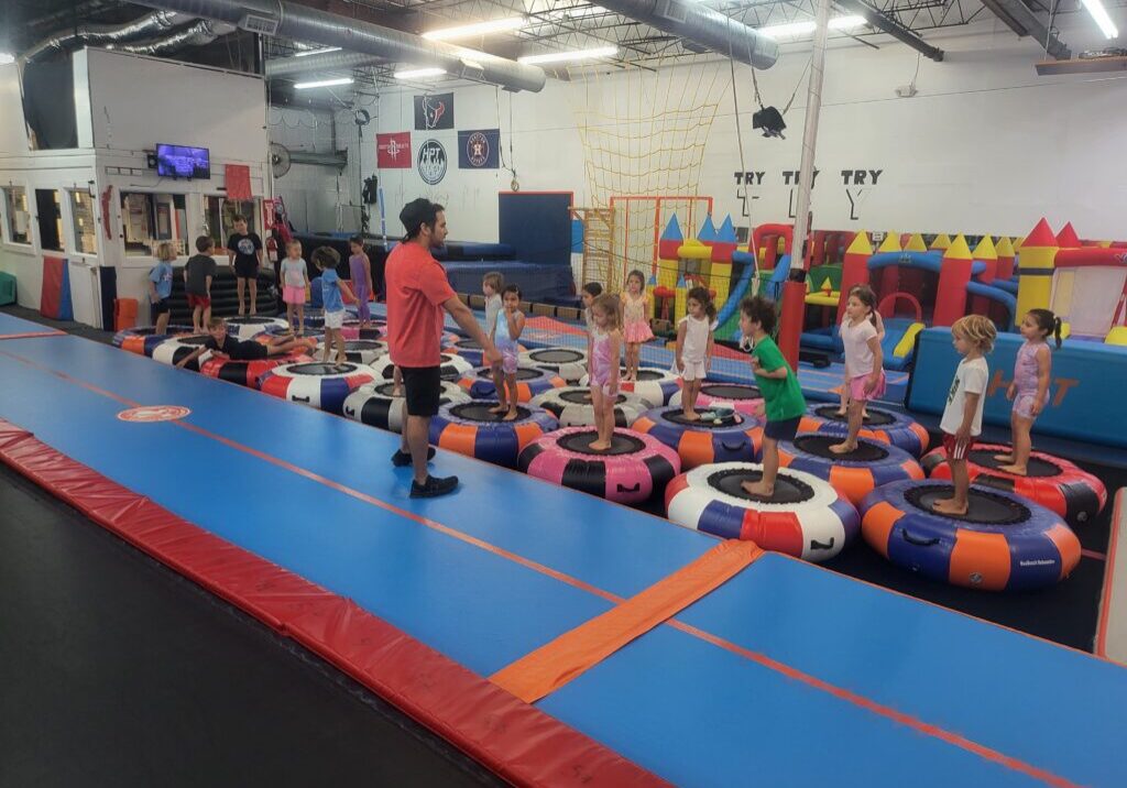 Children stand on trampolines in a gym, guided by an adult in a red shirt. Colorful equipment surrounds them in a bright space.